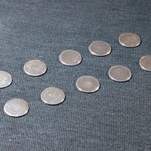 May include: Ten small, circular, metallic discs with a smooth, reflective silver-toned surface. Each disc is flat and uniform in shape, arranged in a scattered pattern across the frame, showcasing a clean, polished appearance without any markings.