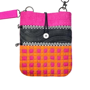 May include: A colorful crossbody bag with a pink strap and accents. The bag features a quilted design with a pink top, black center, and orange and pink checkered bottom. It has a black button closure and silver zippers.