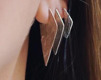 May include: A close-up of a person's ear with three silver triangle earrings. The earrings are layered and have a textured surface.