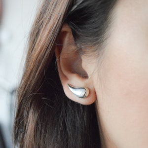 May include: A close-up of a person's ear with a silver teardrop-shaped earring. The earring is a simple, modern design.