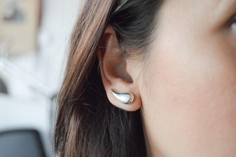 May include: A close-up of a person's ear with a silver teardrop-shaped earring. The earring is a simple, modern design.