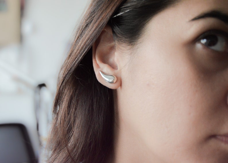 May include: A close-up of a person's ear with a silver teardrop-shaped earring. The earring is a simple, modern design.