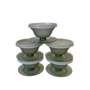 May include: A stack of six vintage green-tinted glass dessert bowls with clear lids. Each bowl sits on a matching saucer. The bowls have a pedestal base and a rounded shape. The lids are a translucent white.