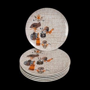 May include: A stack of five round dinner plates with a beige textured background. Each plate features a central graphic print in orange, brown, and black, depicting a rooster, a wooden butter churn, a coffee grinder, and a floral motif.