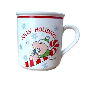May include: White ceramic mug with a red rim and handle. The mug features a cartoon illustration of a character riding a candy cane, with the words "JOLLY HOLIDAYS" and snowflakes. The mug is a festive holiday item.