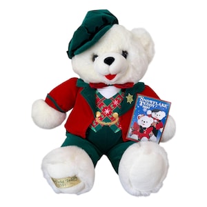 May include: A white plush teddy bear wearing a green hat, red jacket, green vest, and green pants. The bear has a red bow tie and is holding a small book that says "Snowflake Teddy 1997". The bear has a gold tag on its foot that says "Snowflake Teddy 1997".
