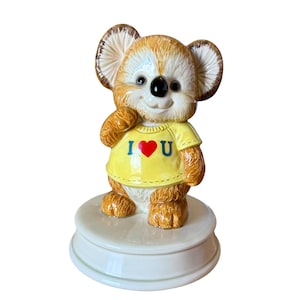 Puede incluir: Figura de koala de cerámica con una camiseta amarilla que dice "I ❤️ U". El koala tiene pelaje marrón, orejas grandes y una nariz negra. Se encuentra sobre una base redonda blanca.