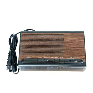 Puede incluir: Un despertador digital vintage con acabado de vetas de madera. El reloj tiene forma rectangular con una rejilla de altavoz y una serie de botones etiquetados como "Snooze", "Hour", "Minute", "Time", "Wake" y "Sleep". Un cable de alimentaci&oacute;n est&aacute; conectado.