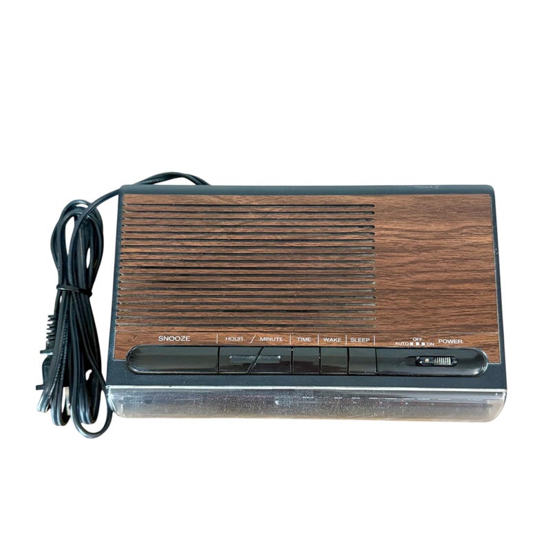 Puede incluir: Un despertador digital vintage con acabado de vetas de madera. El reloj tiene forma rectangular con una rejilla de altavoz y una serie de botones etiquetados como "Snooze", "Hour", "Minute", "Time", "Wake" y "Sleep". Un cable de alimentaci&oacute;n est&aacute; conectado.