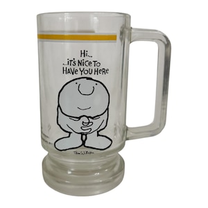 May include: Clear glass beer mug with a handle, featuring a cartoon character illustration and the text "Hi... It's Nice To Have You Here". The mug has a yellow stripe near the top and a wide base.