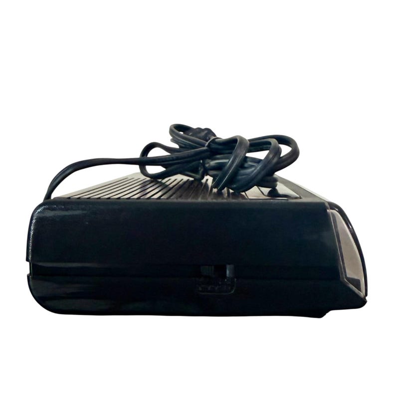 Puede incluir: Un despertador negro de estilo retro con una parte superior acanalada y un cable de alimentaci&oacute;n. El reloj tiene forma rectangular con bordes redondeados y un peque&ntilde;o interruptor lateral. El cable de alimentaci&oacute;n es negro y est&aacute; enrollado encima del reloj.