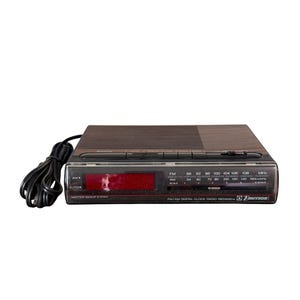 Puede incluir: Un radio reloj digital FM/AM Emerson vintage. La radio tiene una parte superior marr&oacute;n oscuro con un panel frontal negro. La pantalla muestra d&iacute;gitos rojos. La radio tiene un cable de alimentaci&oacute;n negro y escalas de radio AM/FM.