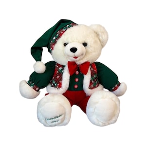 May include: A white plush teddy bear wearing a green and red Christmas outfit. The bear has a green hat, a red bow tie, and a vest with a red and green plaid pattern. The bear's red pants and white paws have the text "Snowflake 1994".