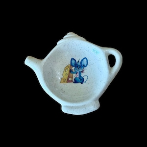 Puede incluir: Un reposacucharas de cerámica blanca con forma de tetera en miniatura y acabado moteado. El centro presenta una colorida ilustración de un ratón azul de pie junto a un trozo de queso suizo amarillo con agujeros visibles en el queso.
