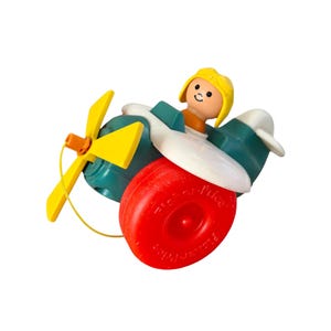 May include: A vintage Fisher-Price toy airplane. The toy features a teal body with white wings, a yellow propeller, and a red wheel. A small figure with a yellow hat and orange shirt sits in the cockpit. The toy is designed for young children.
