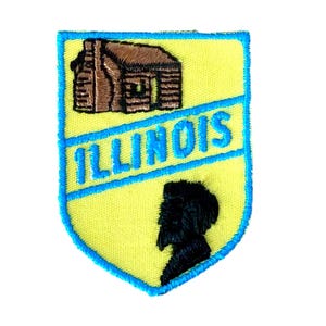 Peut inclure: Un écusson jaune avec une bordure bleue et le mot "ILLINOIS" en bleu. L'écusson représente une cabane marron et une silhouette noire d'une personne.