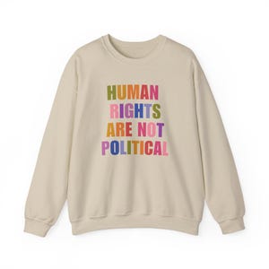 Op de afbeelding: Een beige sweater met ronde hals en de tekst "HUMAN RIGHTS ARE NOT POLITICAL" gedrukt in verschillende kleuren, waaronder groen, rood, roze, blauw, geel en paars. Het kledingstuk heeft lange mouwen en een geribbelde kraag, boorden en zoom.