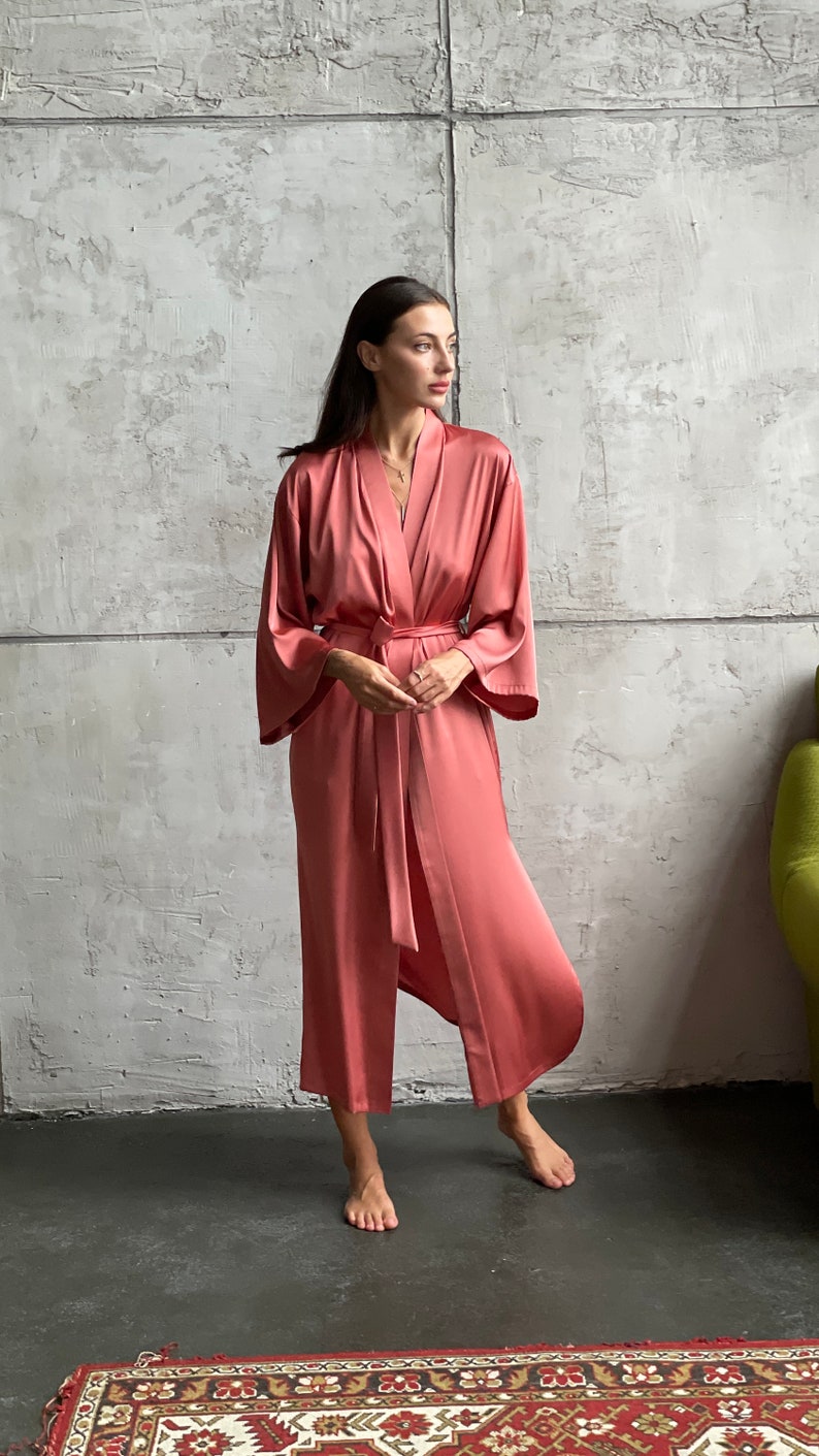 May include: A long, salmon-colored satin robe with a tie belt. The robe has wide sleeves and a relaxed fit.