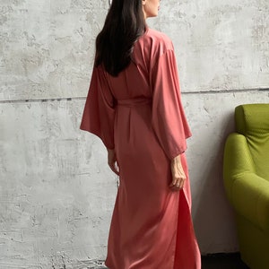 May include: A long, salmon-colored satin robe with a tie belt. The robe has long sleeves and a side slit. The model is wearing the robe and walking away from the camera.