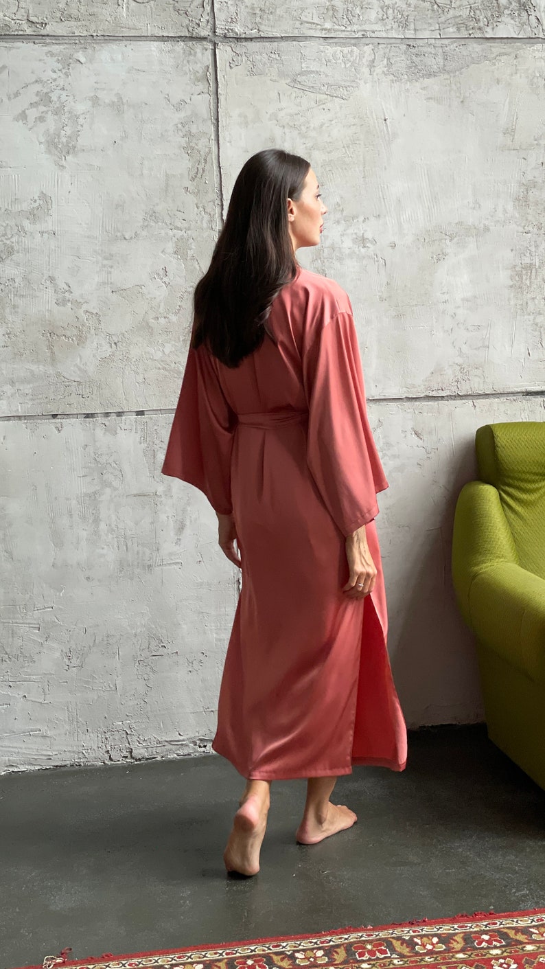 May include: A long, salmon-colored satin robe with a tie belt. The robe has long sleeves and a side slit. The model is wearing the robe and walking away from the camera.