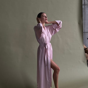 May include: A woman wearing a long, light pink robe with a sash tied at the waist. She is standing on a green background and looking up.
