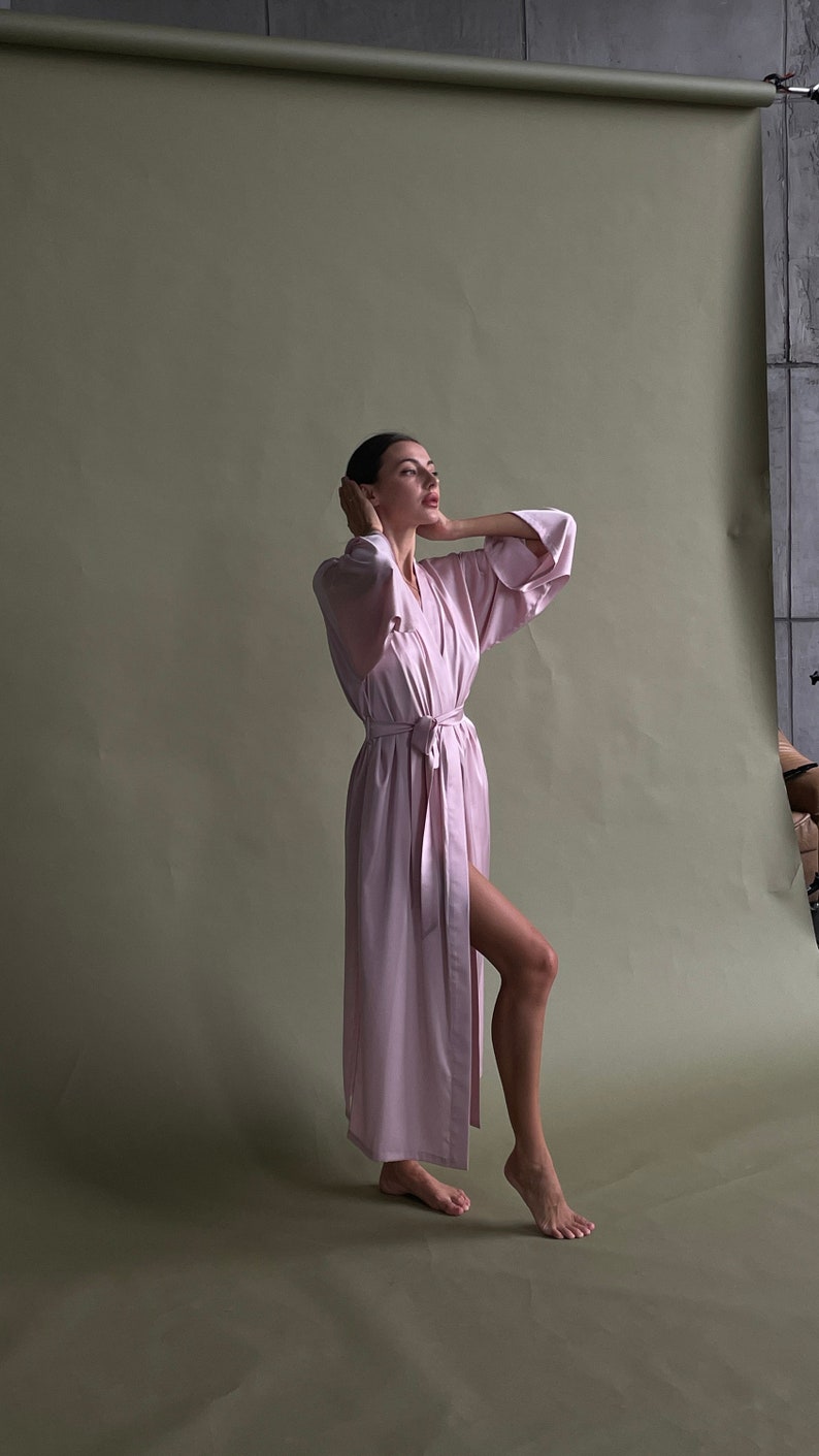 May include: A woman wearing a long, light pink robe with a sash tied at the waist. She is standing on a green background and looking up.
