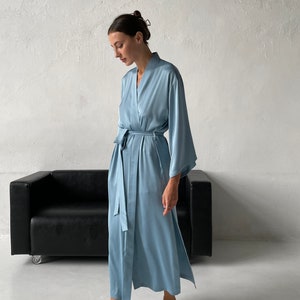 May include: A light blue satin robe with long sleeves and a tie belt. The robe is worn by a person standing in front of a white wall and a black couch.