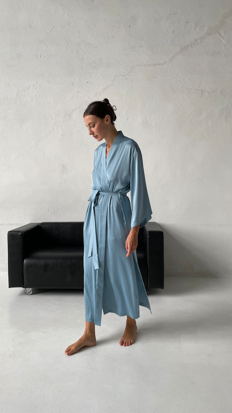 May include: A light blue satin robe with long sleeves and a tie belt. The robe is worn by a person standing in front of a white wall and a black couch.