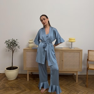 Könnte beinhalten: Hellblaues Satin-Pyjama-Set, bestehend aus einem Morgenmantel mit ausgestellten Ärmeln, einem Camisole und einer Hose mit Rüschensaum. Das Set wird mit einem passenden Gürtel in der Taille gebunden. Das Outfit wird in Innenräumen getragen.