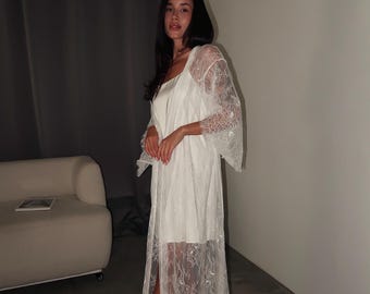 Puede incluir: Una bata de encaje blanco hasta el suelo con mangas anchas de tres cuartos y diseño frontal abierto. La prenda está confeccionada con un delicado encaje floral semitransparente que llega hasta el dobladillo, sobre un vestido lencero blanco liso.