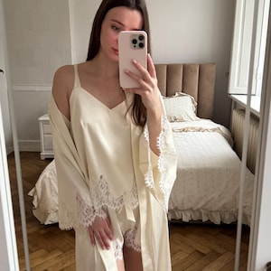 Peut inclure: Ensemble de pyjama en satin ivoire avec bordure en dentelle. L'ensemble comprend un caraco, un short et une longue robe. La robe a des manches larges et une fermeture à nouer. Chambre avec lit et tête de lit beige en arrière-plan.