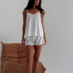 May include: White camisole top and matching shorts. The top has thin straps and a V-neck. The shorts have a drawstring waist. The outfit is made of a smooth, silky fabric. A brown, tufted lounge chair is in the background.