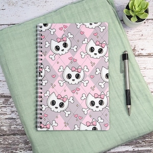 May include: A spiral-bound notebook featuring a repeating pattern of white cat skulls with pink bows and crossbones on a background of light pink and gray diamonds, accented with small pink hearts and four-pointed star shapes throughout the design.