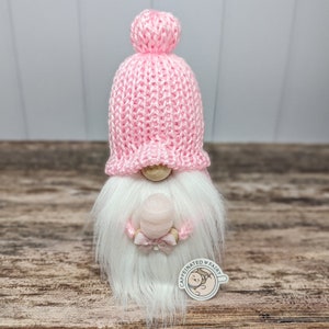 May include: A pink knitted gnome with a white beard and a pink pom-pom hat. The gnome is holding a pink ball with a pink ribbon. The gnome is sitting on a wooden surface. The text "Caffeinated Fairy" is on a sticker on the gnome's leg.