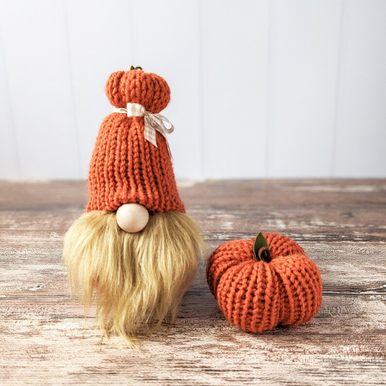 May include: A knitted gnome wearing a pumpkin hat with a white and brown ribbon. The gnome is sitting on a wooden surface next to a knitted pumpkin.