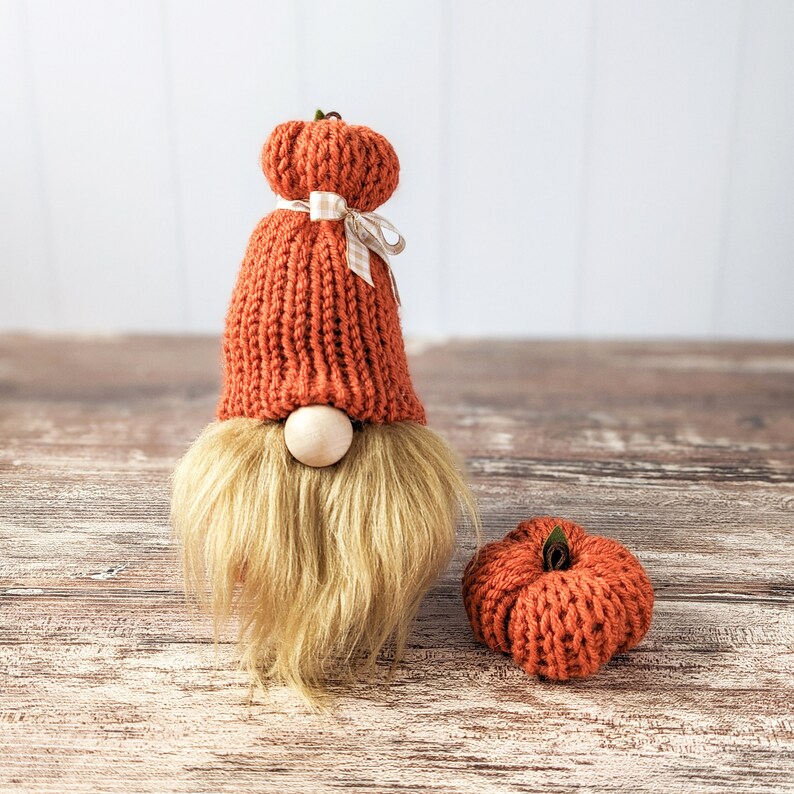 May include: A knitted gnome with a red and white hat and a brown beard. The gnome is holding a small knitted pumpkin in its hand.