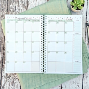 May include: A spiral-bound monthly planner open to August 2027, featuring a white grid layout with light green headers for days of the week. The top includes pink star icons and a small cat illustration. The right side has sections for goals, to-do lists, and notes.