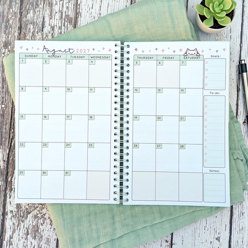 May include: A spiral-bound monthly planner open to August 2027, featuring a white grid layout with light green headers for days of the week. The top includes pink star icons and a small cat illustration. The right side has sections for goals, to-do lists, and notes.
