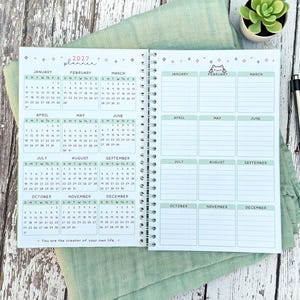 May include: A spiral-bound 2027 planner featuring a full-year calendar on the left page and a monthly goal or note section on the right page. The pages include light green headers, small star icons, a cat illustration, and the text You are the creator of your own life.