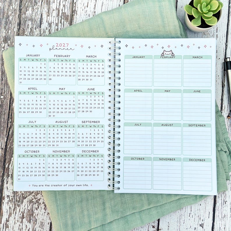 May include: A spiral-bound 2027 planner featuring a full-year calendar on the left page and a monthly goal or note section on the right page. The pages include light green headers, small star icons, a cat illustration, and the text You are the creator of your own life.