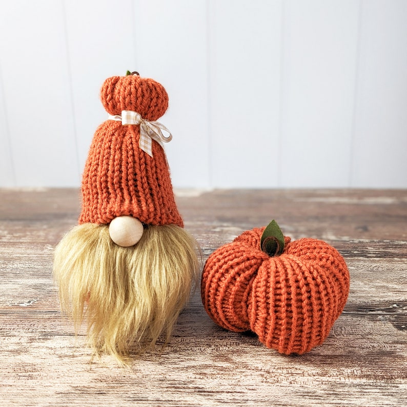 May include: A knitted gnome wearing an orange hat with a white and brown ribbon. The gnome is standing next to a knitted orange pumpkin.