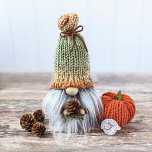 May include: A knitted gnome with a green and orange hat, a white beard, and a brown bow. The gnome is holding a small pine cone in its hand. There is a small orange knitted pumpkin and a sticker with the text "Caffeinated Fairy" on a wooden surface.