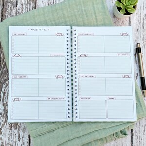 May include: A spiral-bound weekly planner open to a two-page spread for August 16-22. The pages feature daily sections with checkboxes, small cat illustrations, and headers for Sunday through Saturday, plus dedicated sections for priorities and notes.