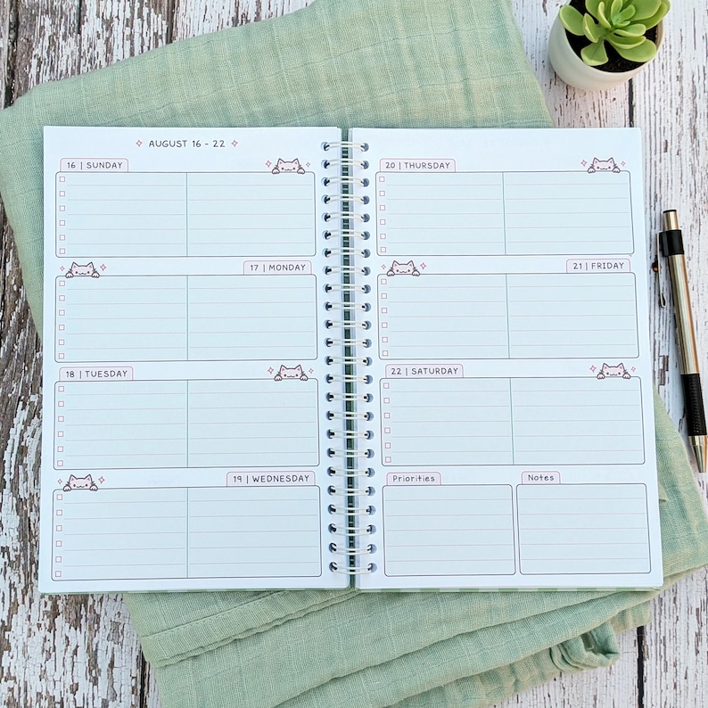 May include: A spiral-bound weekly planner open to a two-page spread for August 16-22. The pages feature daily sections with checkboxes, small cat illustrations, and headers for Sunday through Saturday, plus dedicated sections for priorities and notes.