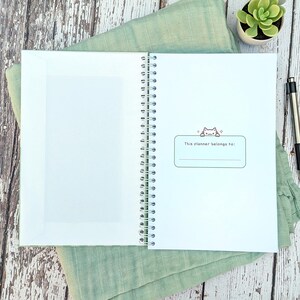 May include: A spiral-bound planner with a white interior page featuring a rectangular label with a small cat illustration and the text "This planner belongs to:" above a blank line. The left side of the planner includes a white paper pocket.