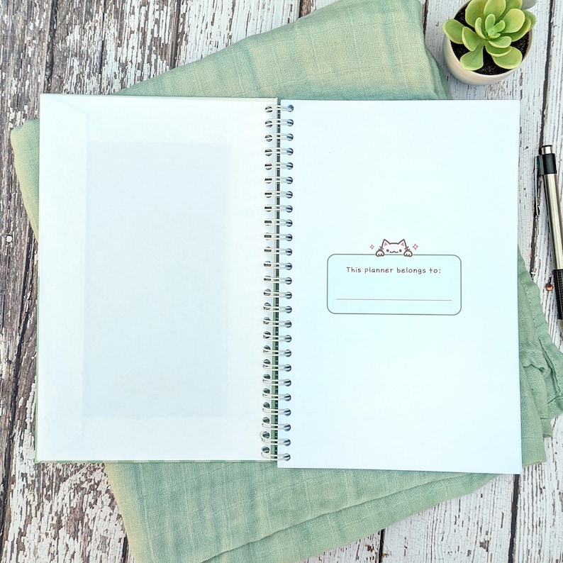 May include: A spiral-bound planner with a white interior page featuring a rectangular label with a small cat illustration and the text "This planner belongs to:" above a blank line. The left side of the planner includes a white paper pocket.