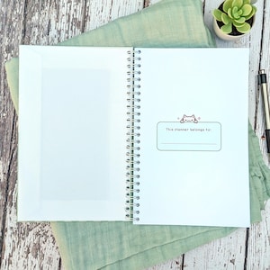 May include: A spiral-bound planner with a white interior page featuring a rectangular label with a small cat illustration and the text "This planner belongs to:" above a blank line. The left side of the planner includes a white paper pocket.
