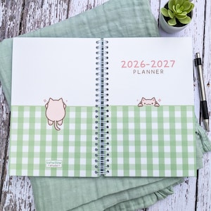 May include: A spiral-bound 2026-2027 planner with a white and light green gingham pattern on the bottom half of the pages. The planner features illustrations of a small pink cat and the text 2026-2027 PLANNER and catwillow studio.