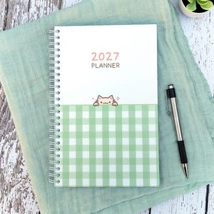 May include: A 2027 spiral-bound planner featuring a white top section with the text 2027 PLANNER and a green and white gingham bottom section. A small cartoon cat illustration sits at the border between the two sections. A black and silver pen.