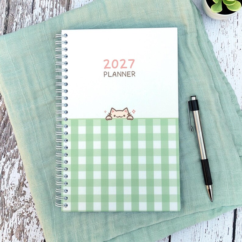 May include: A 2027 spiral-bound planner featuring a white top section with the text 2027 PLANNER and a green and white gingham bottom section. A small cartoon cat illustration sits at the border between the two sections. A black and silver pen.